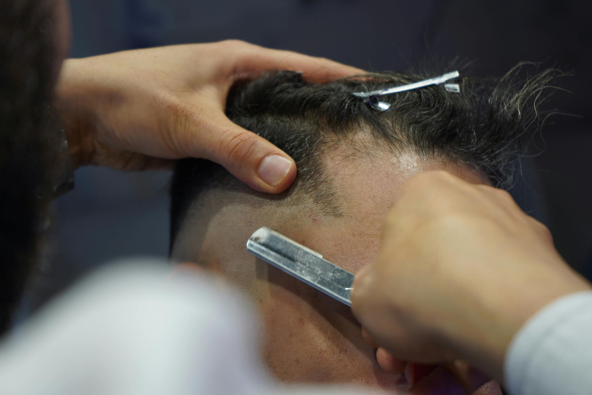 A barber uses a straight razor to edge a client's hairline, holding the hair back with a metal clip.