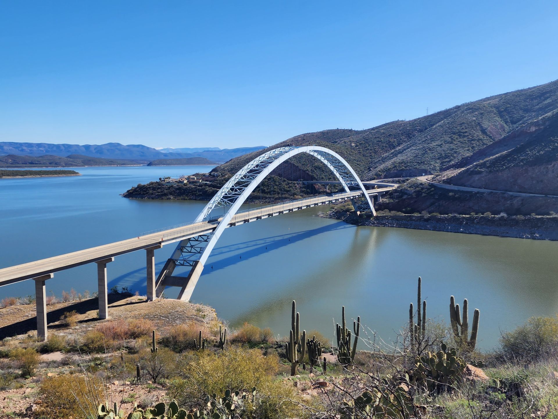 White arch bridge over a calm blue bay beside rocky hills and cactus in the foreground