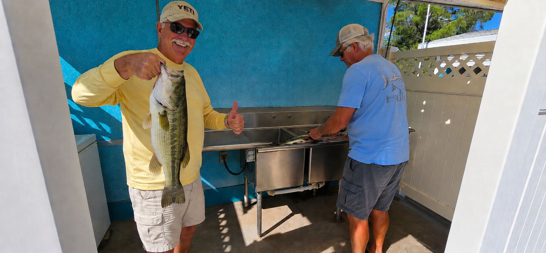 Two men at an outdoor sink under a blue canopy, one holding up a fish while the other rinses a large pot.