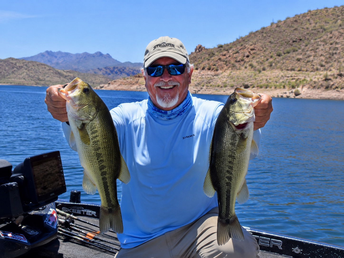 Man holding two large fish on a boat with blue water and desert mountains in the background