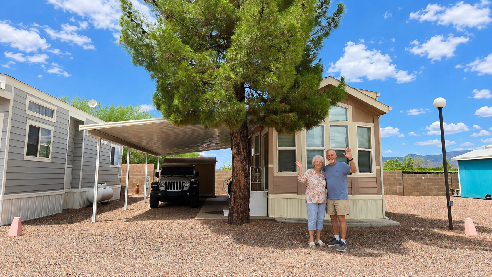 Two people standing beside a small house and carport under a large tree on a sunny day.