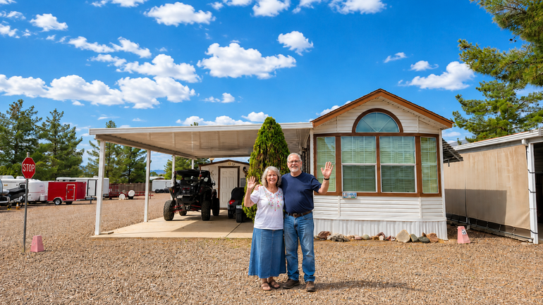 Two people standing outside a small house under a carport on a sunny day.
