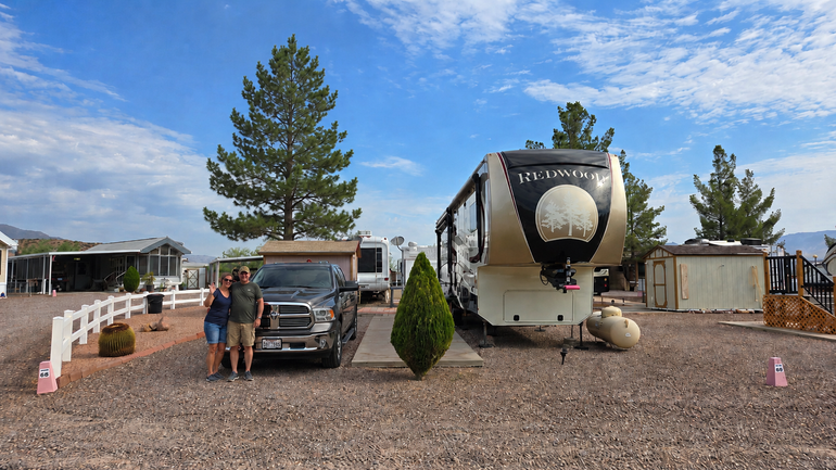 RV and truck parked on gravel at a campsite, with two people standing nearby under a blue sky.