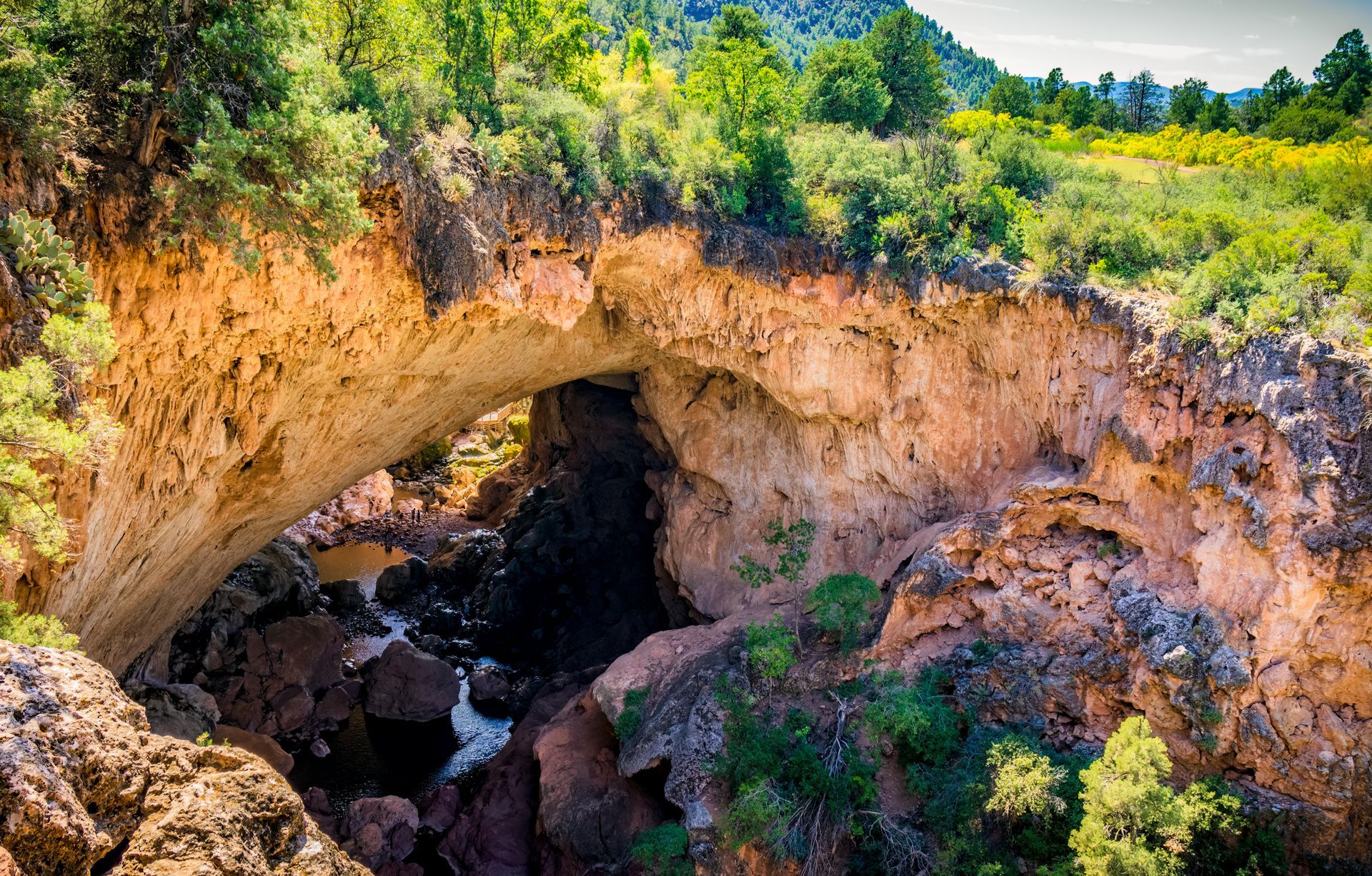 Rock arch over a rocky gorge with a stream, surrounded by green trees and sunlit cliffs