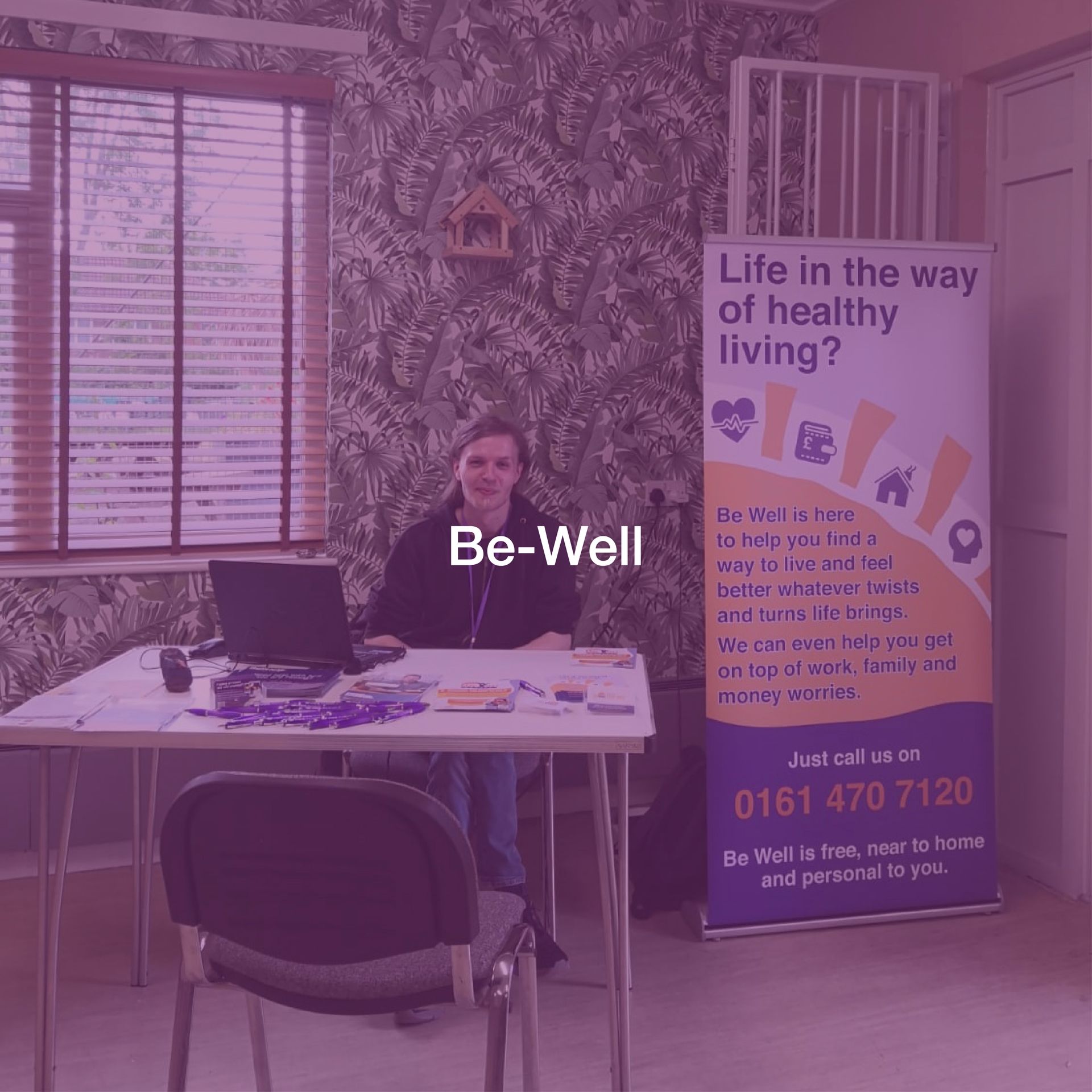 A woman is sitting at a desk in front of a sign that says be well