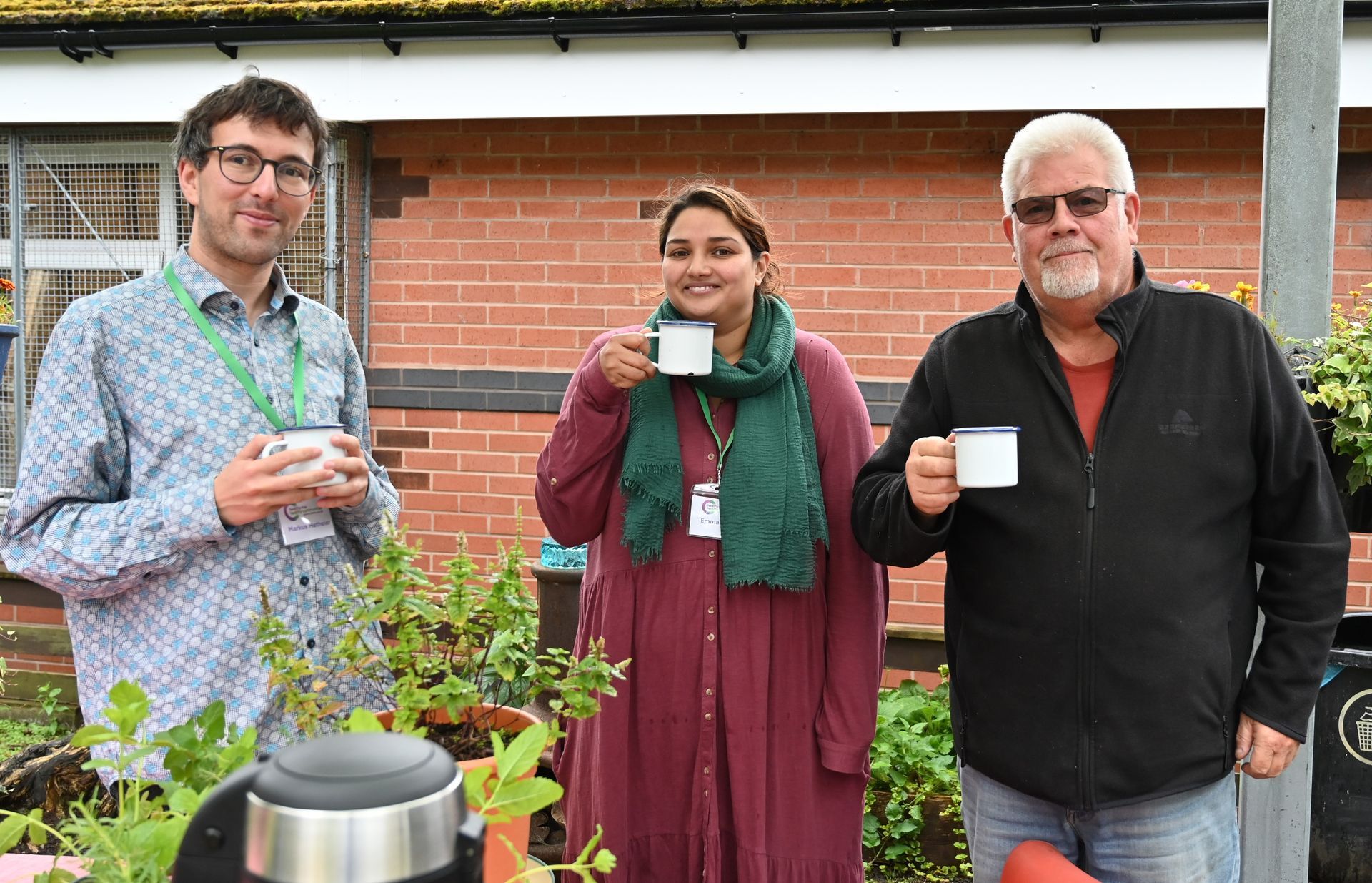 Three people are standing next to each other, holding cups of coffee. They are out in the garden. There is a red brick building behind them and plants in pots in front of them. 