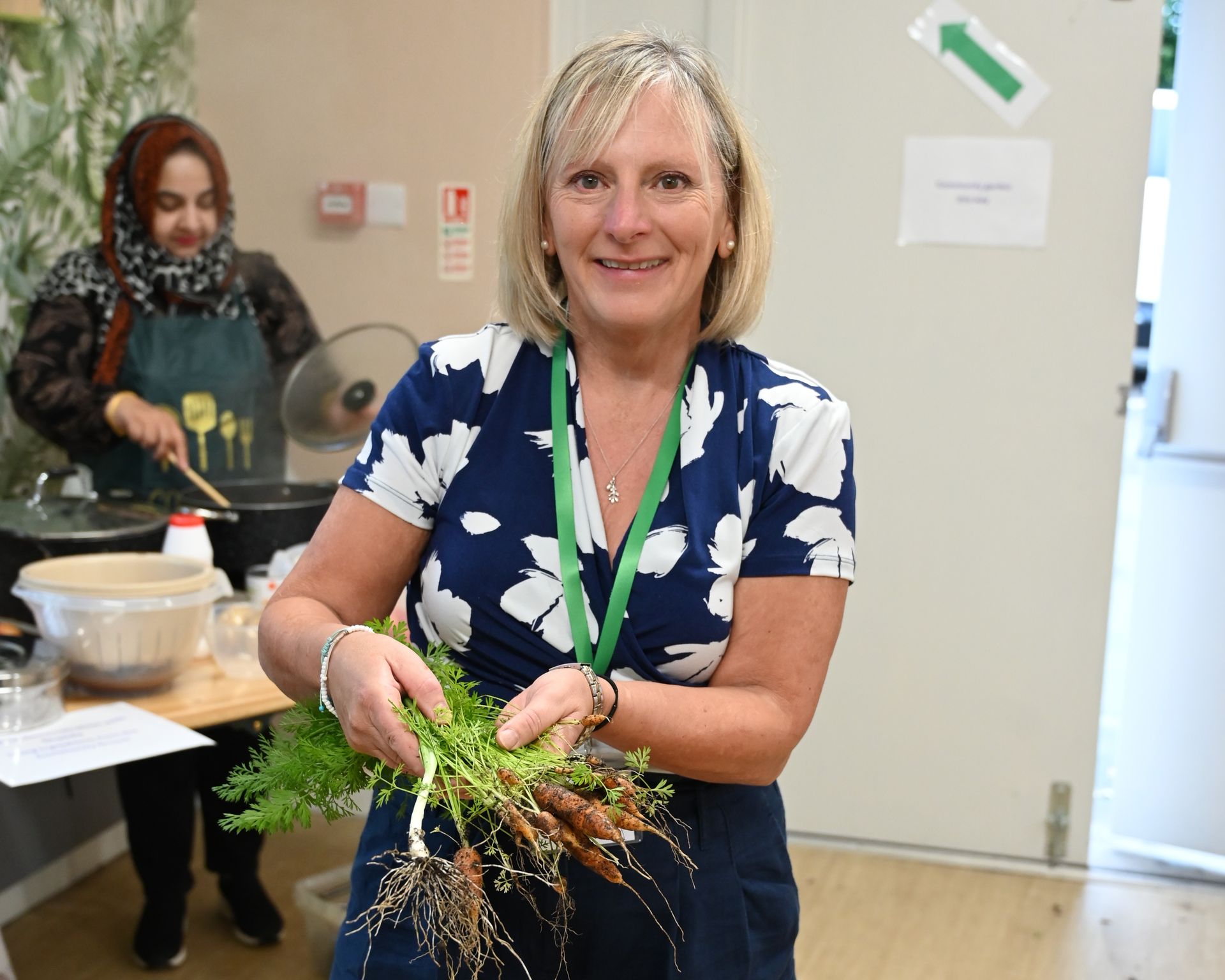 A woman is holding a bunch of carrots in her hands. Behind her another woman is cooking in a pan.