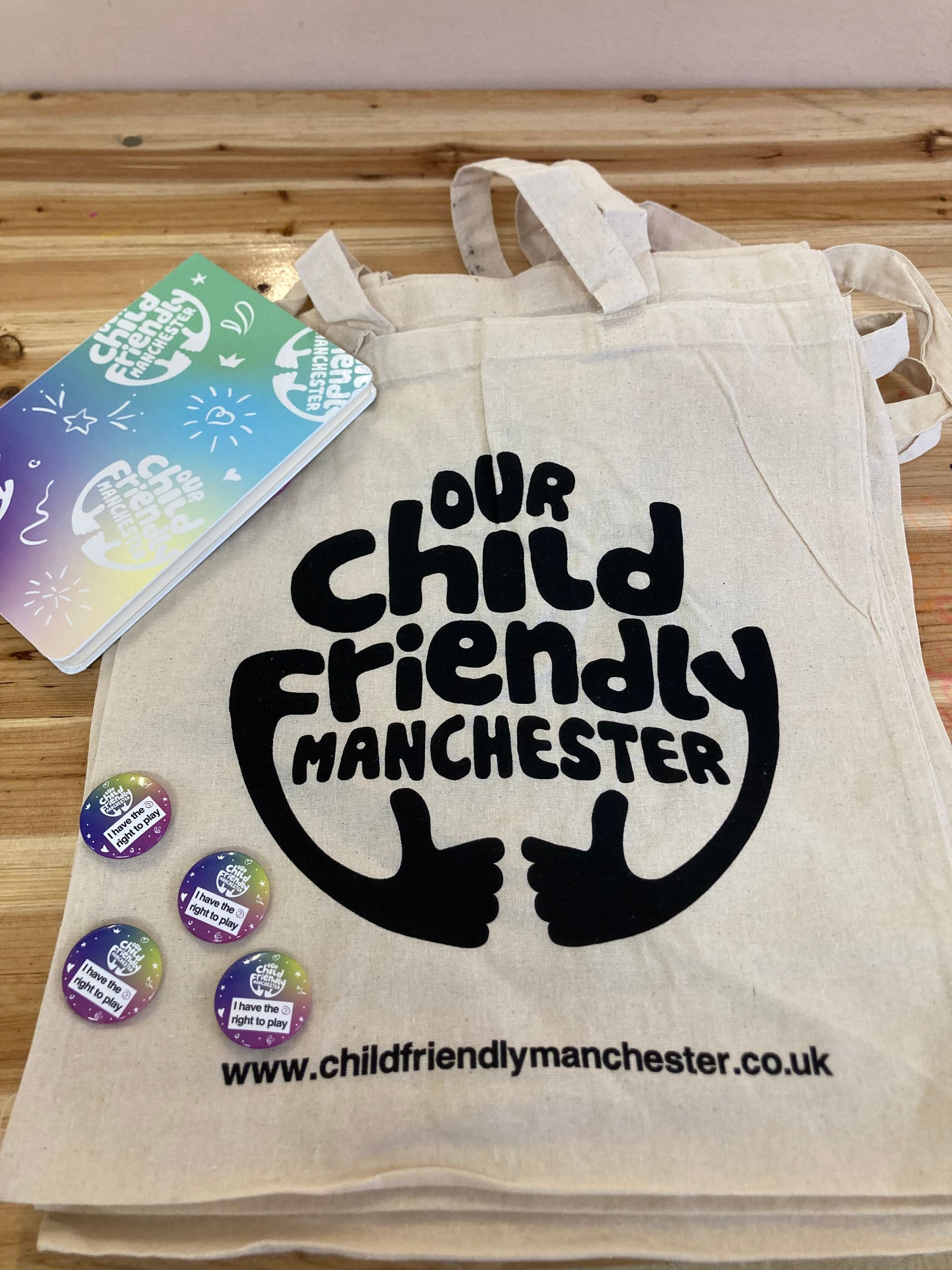 White cloth tote bag with the logo of Our Child Friendly Manchester in large print. The bag is on a table with colourful pin badges and a notebook.  