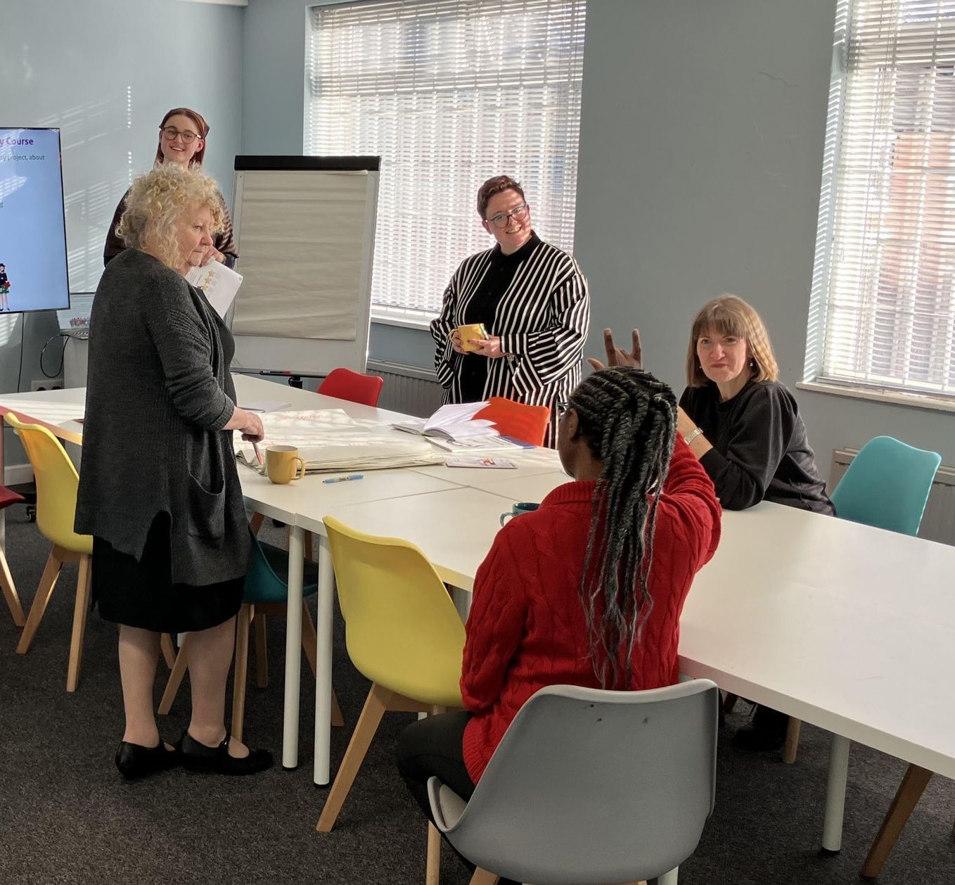 A group of women are sitting around a table in a room. The room is modern and bright with natural light. There is a flipchart stand in the background. 