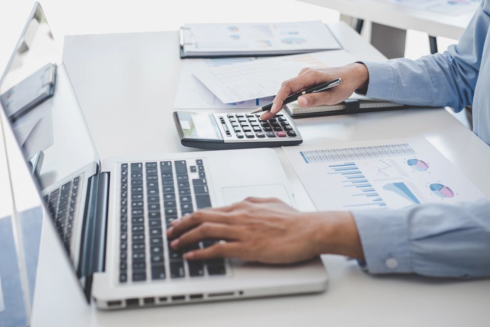 Persona que usa una calculadora y una computadora portátil, trabajando con datos financieros en un escritorio.
