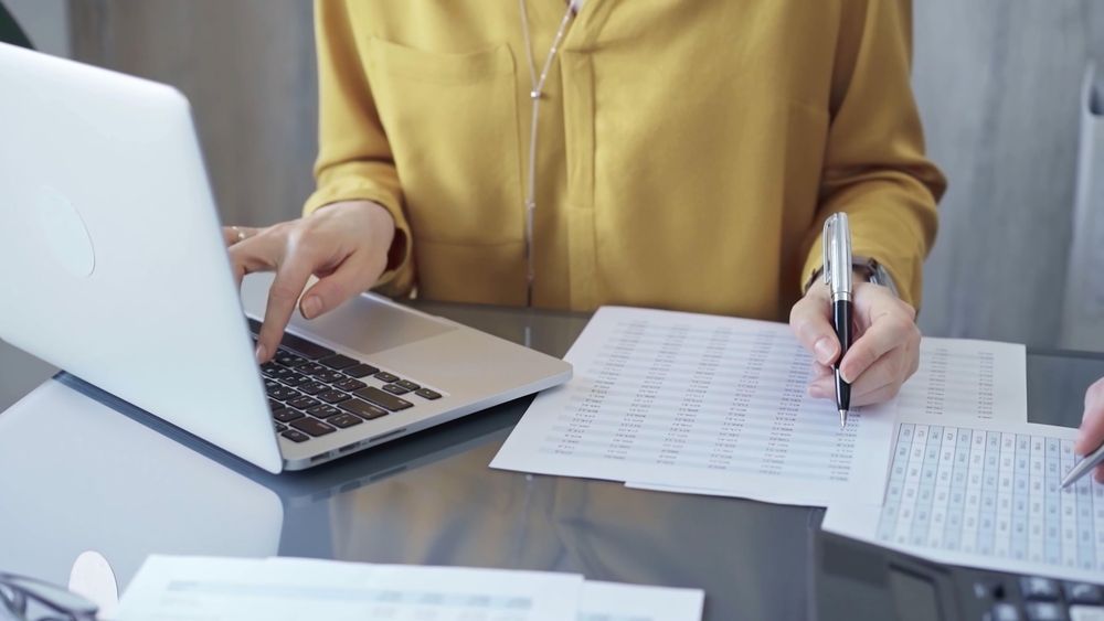 Persona con blusa amarilla, escribiendo en una computadora portátil mientras escribe en un documento, en una mesa con papeles.