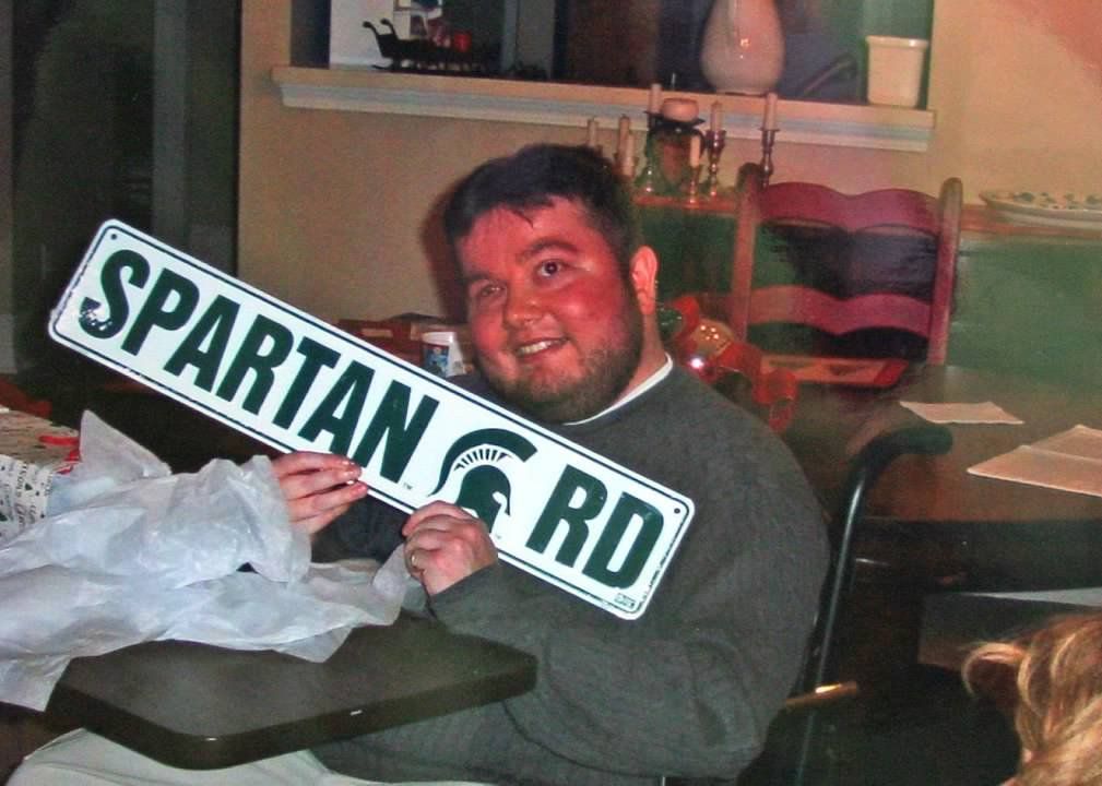 Man holding a large SPARTAN RD street sign indoors, smiling at the camera.