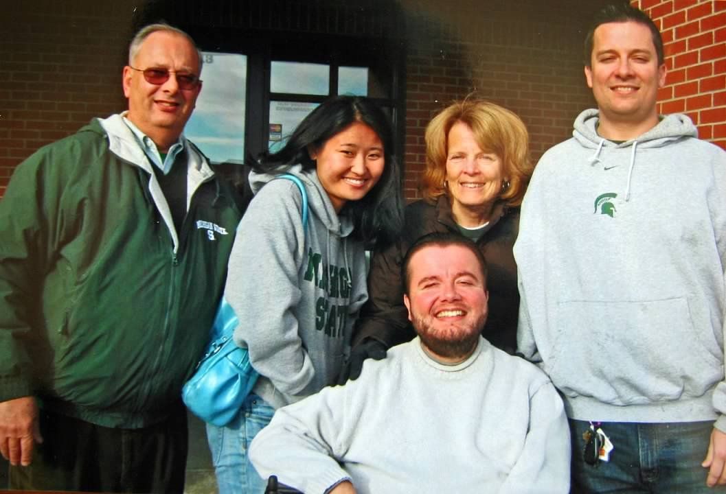 Group of five people smiling outdoors, one in a wheelchair, all wearing casual clothing near a brick building.