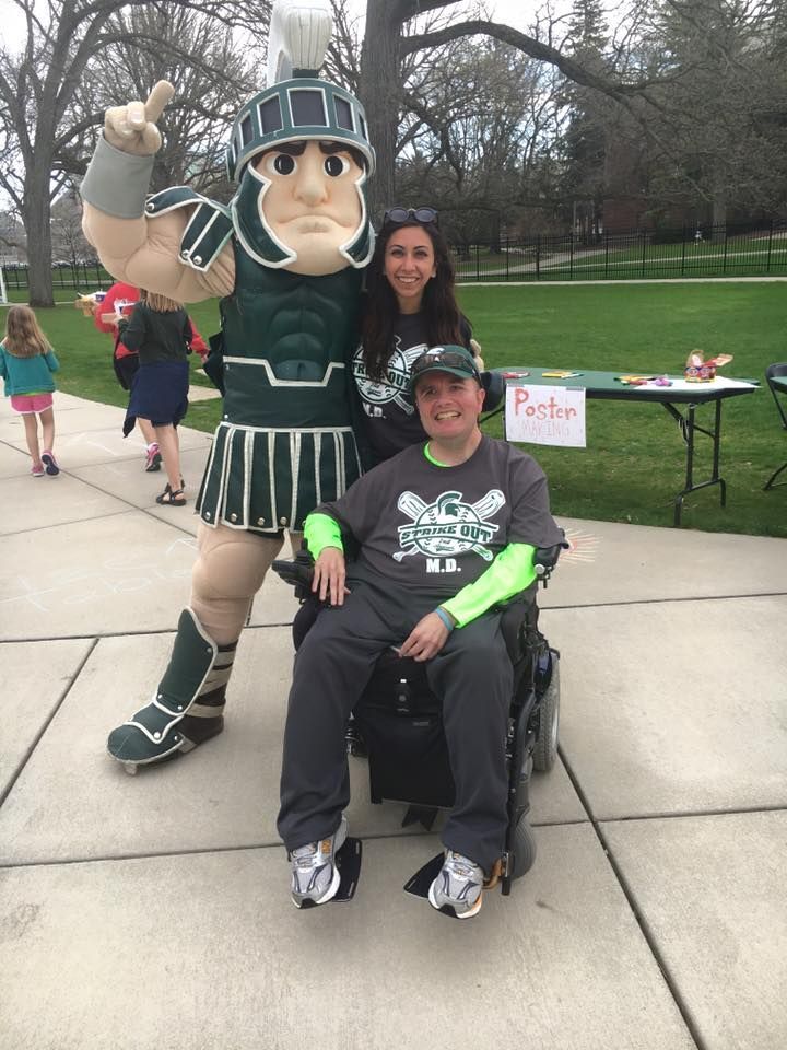 Person in a Spartan mascot costume posing with a person in a wheelchair outdoors near a table and sign.