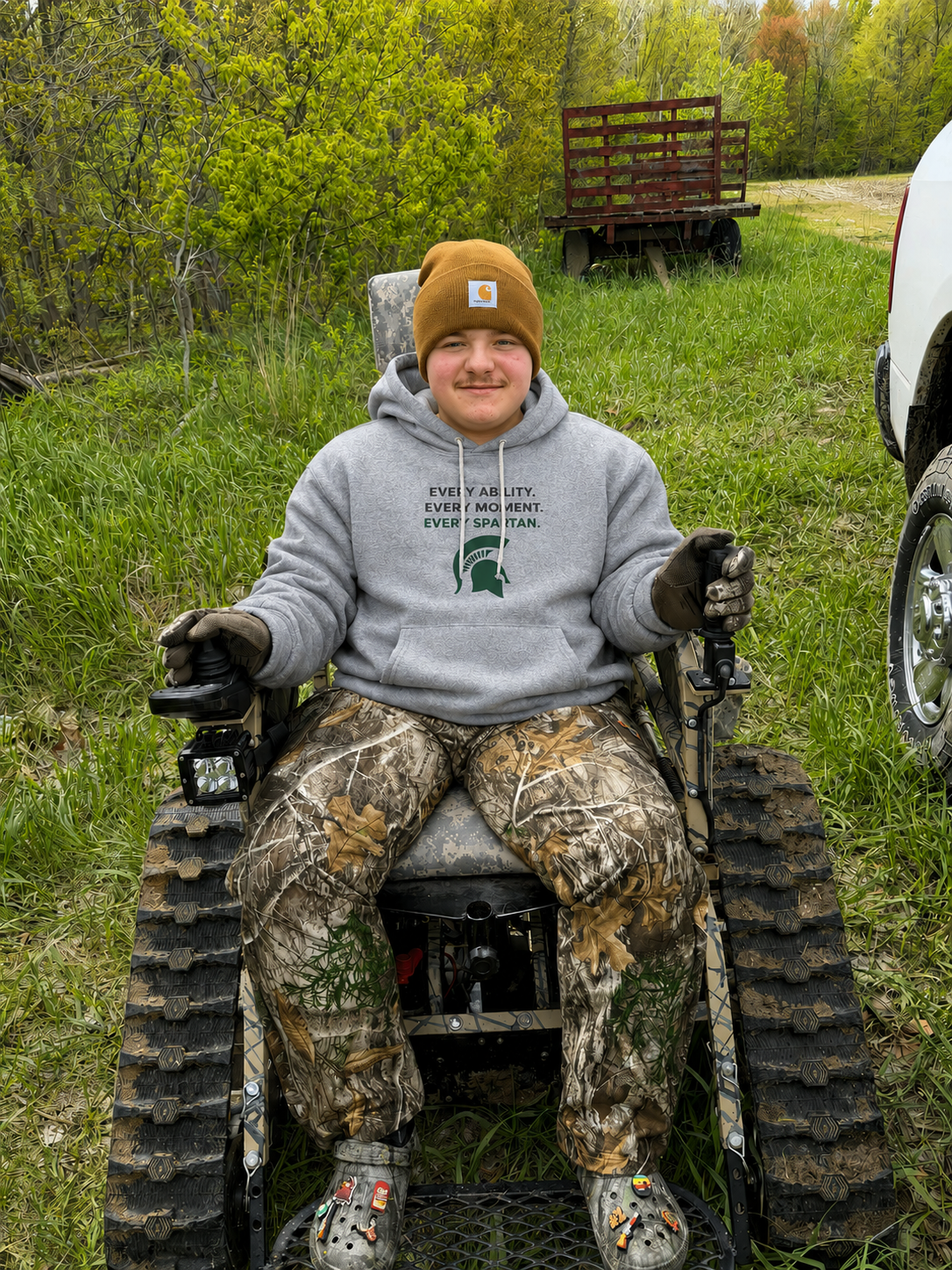 Person smiling in a tracked wheelchair outdoors on a grassy path, wearing a gray hoodie and brown beanie.