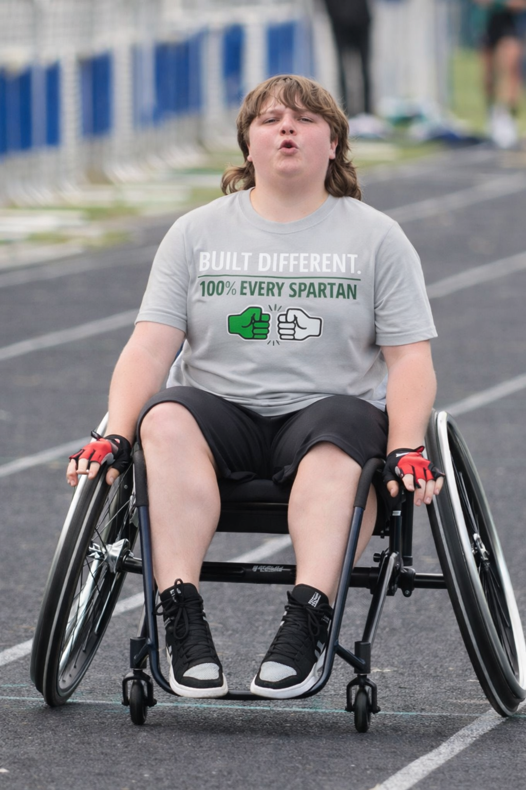 Person in a wheelchair on a road, wearing a gray shirt and black shorts, looking ahead.