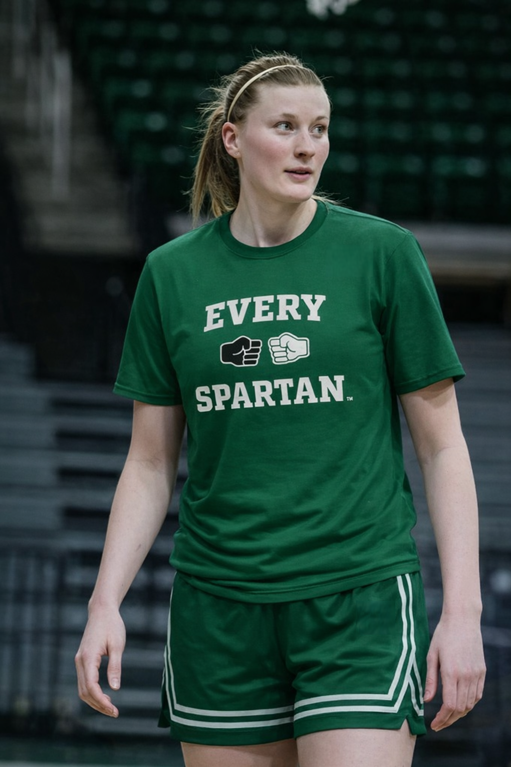 Woman in green shirt and shorts, standing in a basketball arena. Shirt reads 