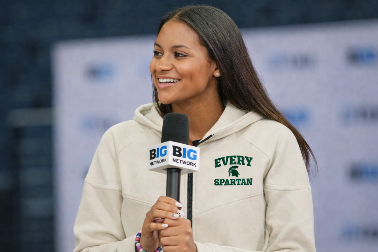 Person smiling and speaking into a microphone at a Big 12 event, wearing a beige hoodie.