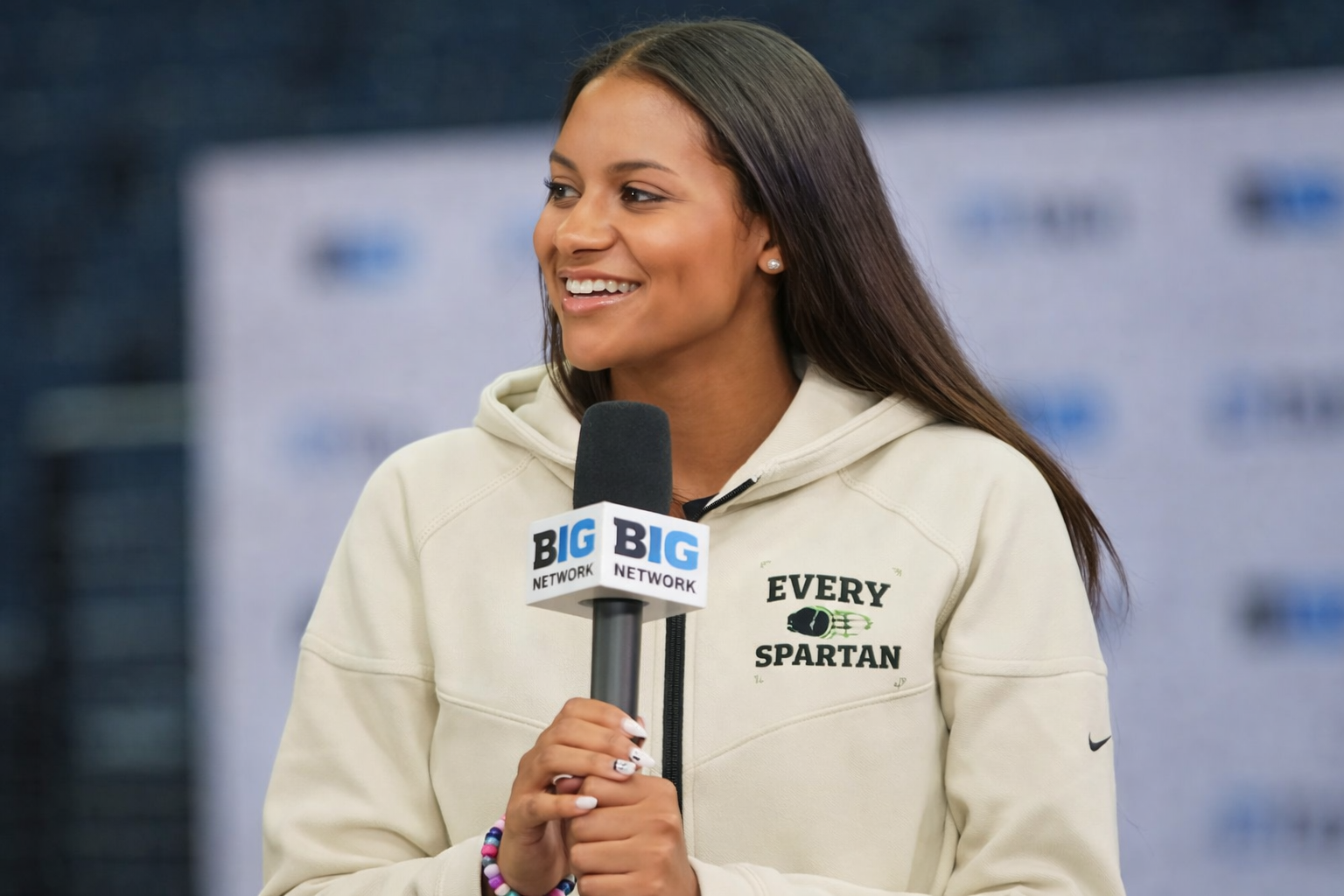 Woman smiling, holding a microphone. She wears a beige hoodie that says 