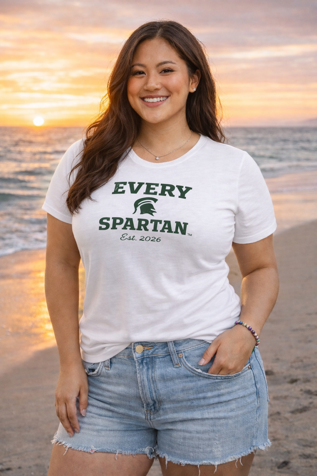 Woman on beach wearing a white t-shirt with green text, denim shorts. Sunset in background.