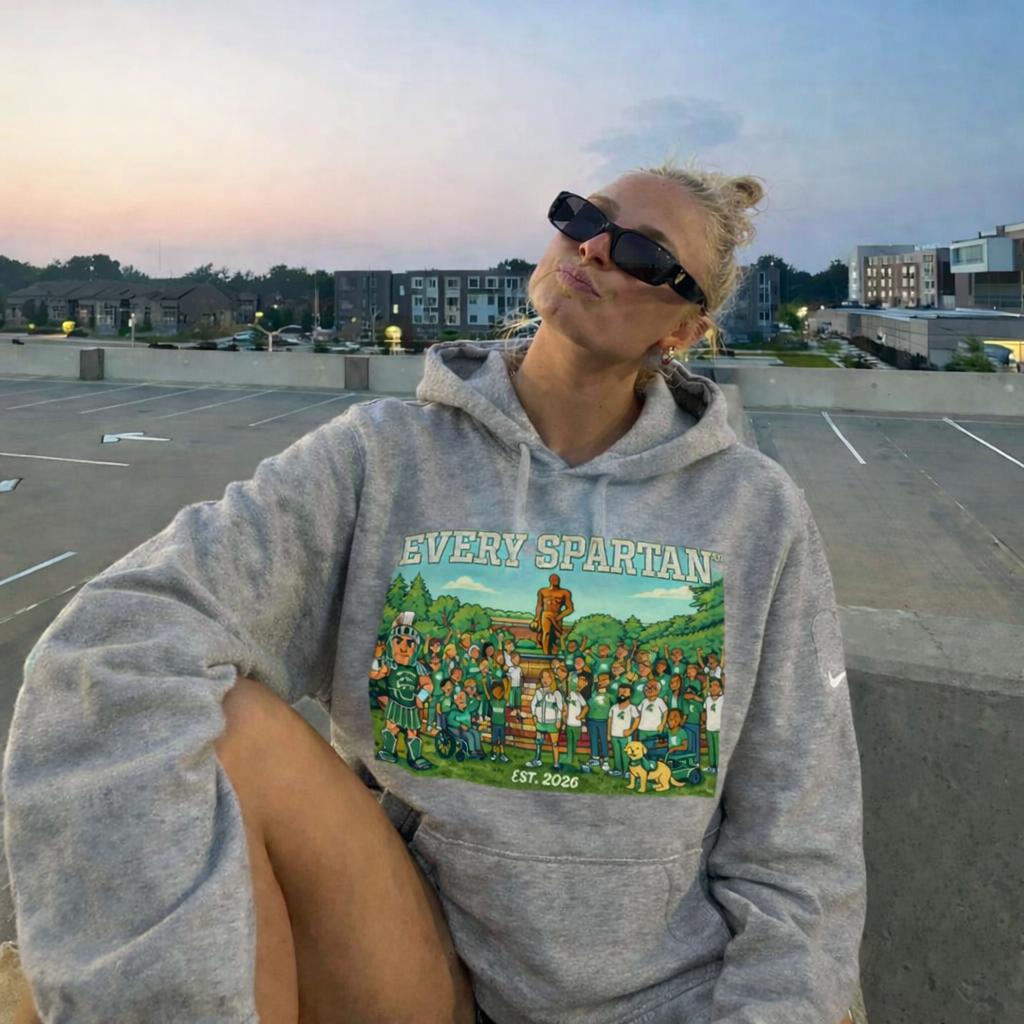 Woman in sunglasses and gray hoodie on a rooftop, puckering lips. Hoodie features a cartoon group and 