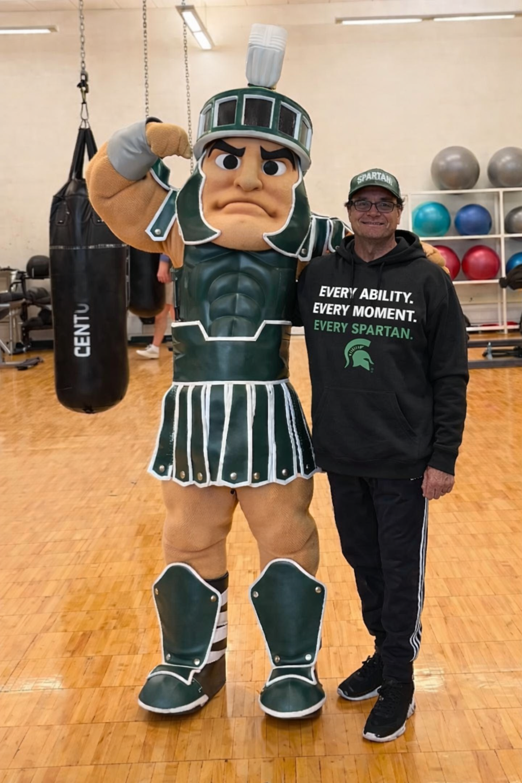 Spartan mascot flexing near a man in a Spartan hoodie, in a gym with exercise equipment.
