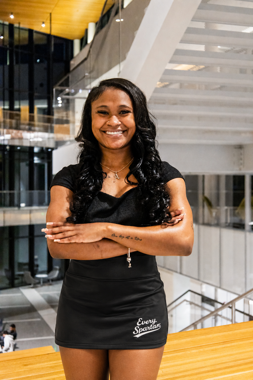 Smiling woman in black dress standing with arms crossed in a modern atrium with stairs and glass railings