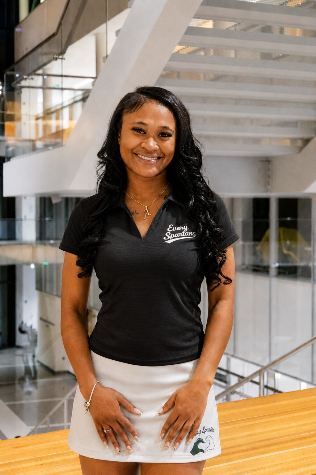 Smiling woman in black polo and white skirt standing in a modern indoor atrium
