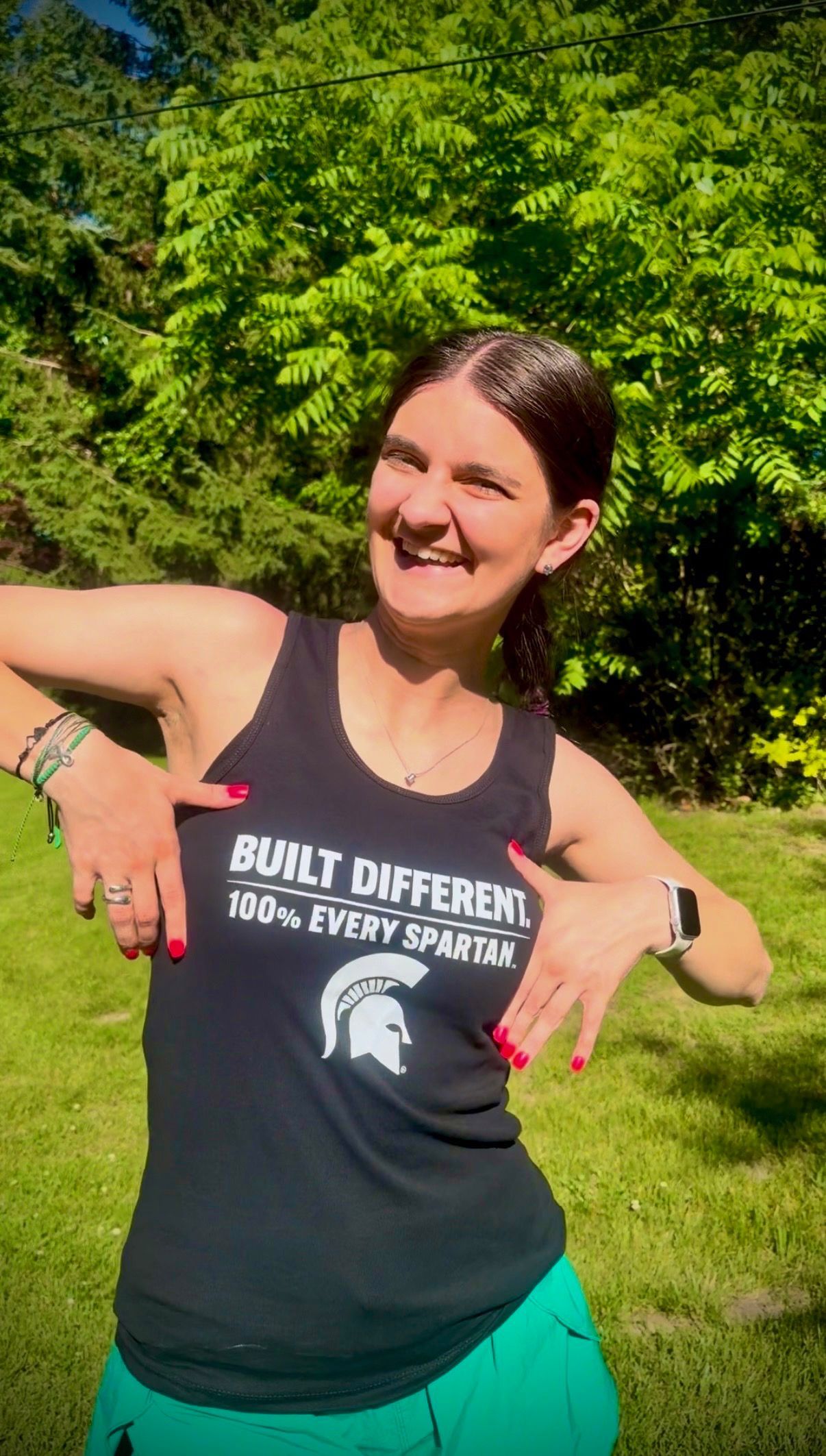 Smiling person outdoors in a black tank top and teal shorts, flexing both arms in a green park.