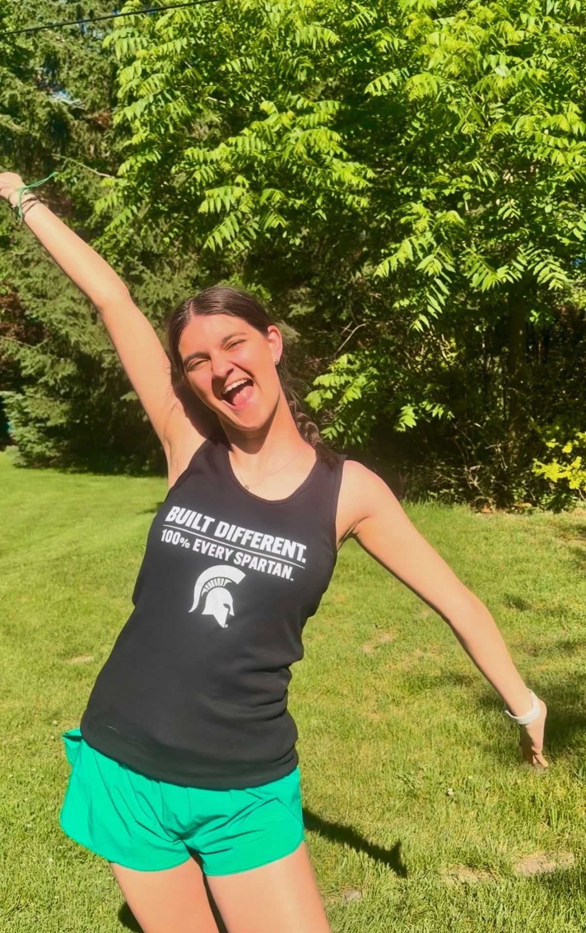 Smiling person in black tank top and green shorts posing with arms raised in a sunny grassy park