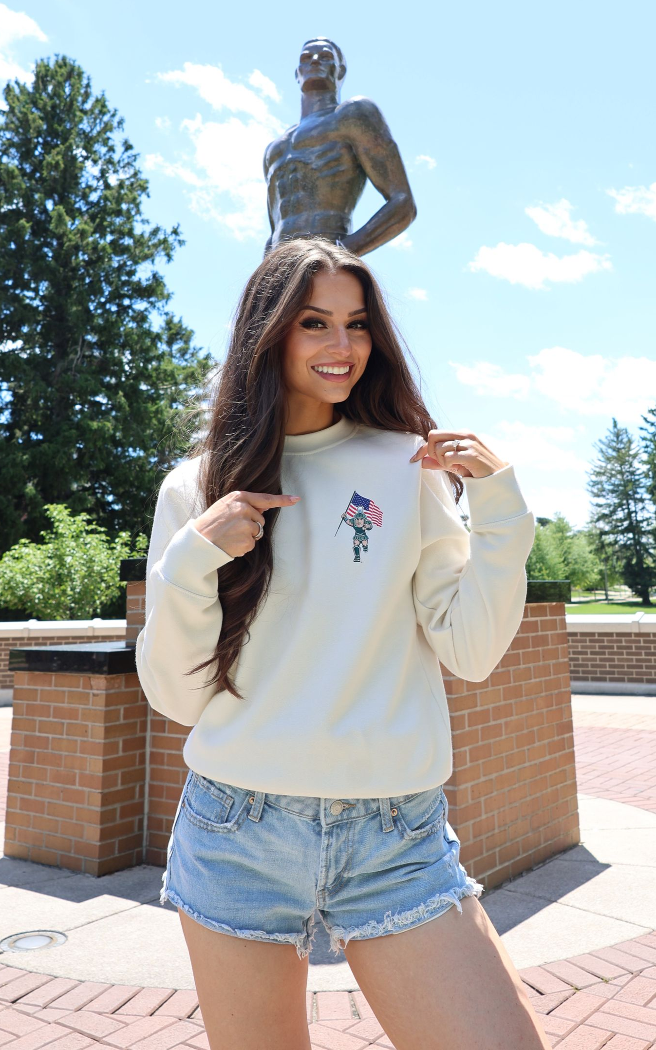 Woman in a white sweatshirt and denim shorts posing outdoors in front of a statue.