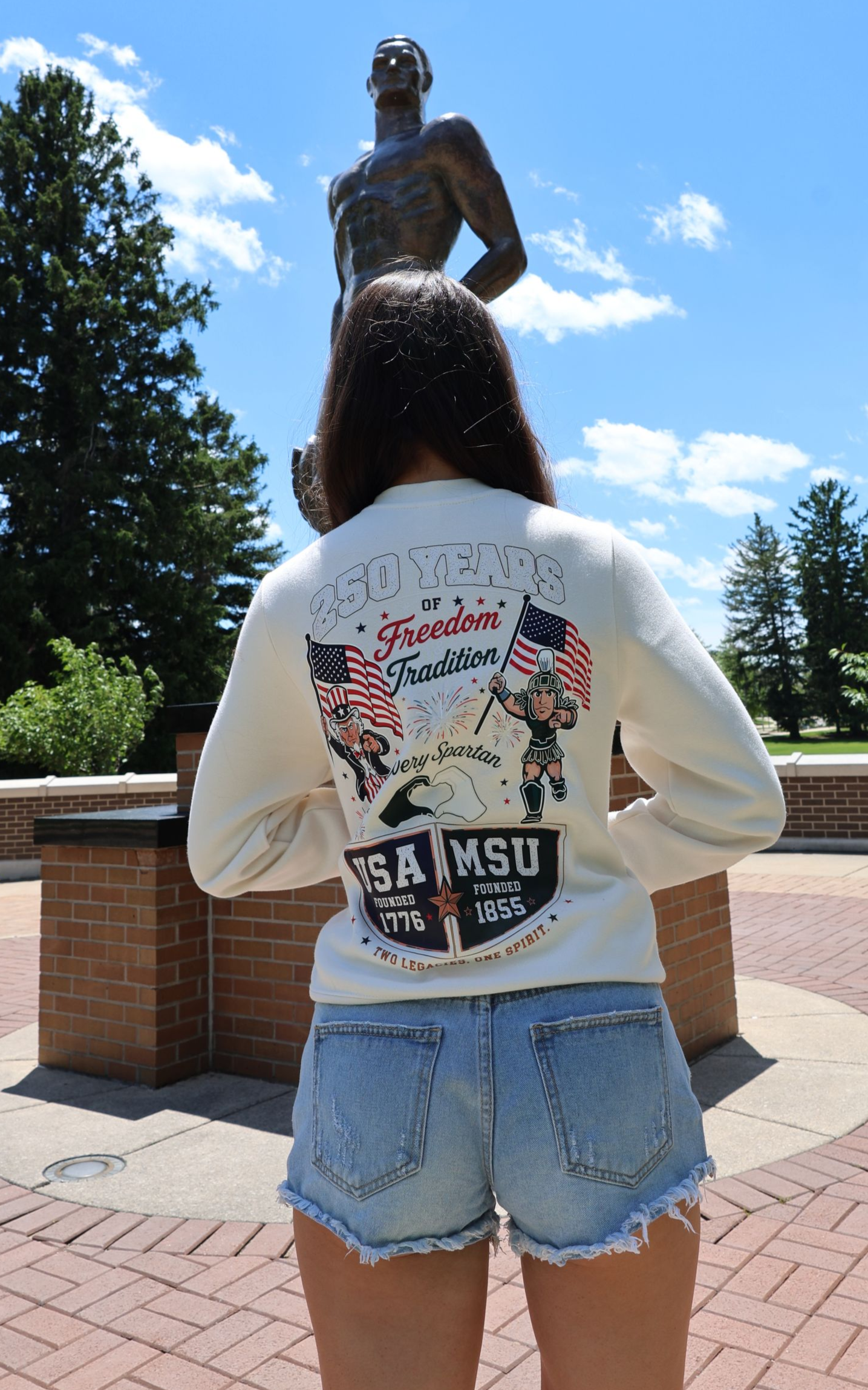 Person in white shirt and denim shorts facing a bronze statue on a sunny brick walkway.