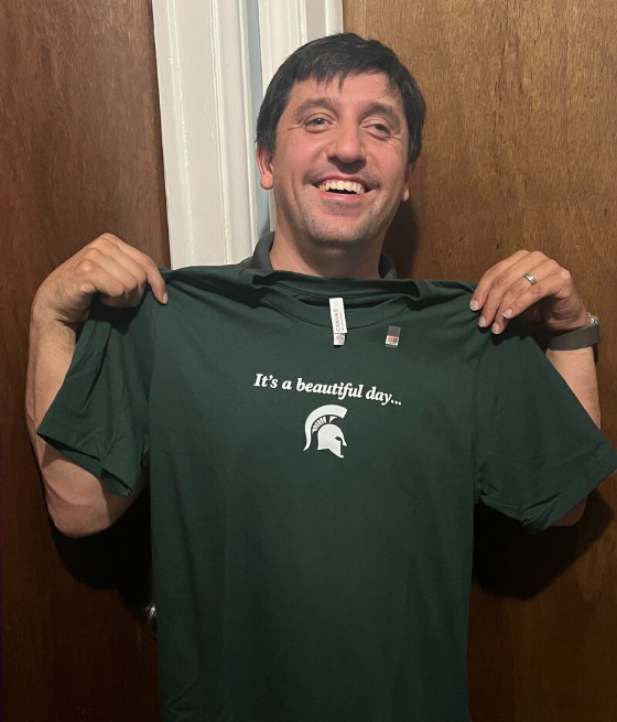 Man smiling and holding up a dark green T-shirt with white text against wooden doors.