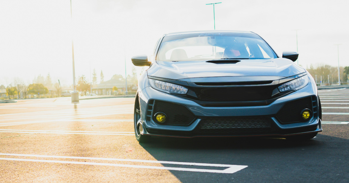 Dark gray car parked on asphalt, yellow fog lights, sunny day.