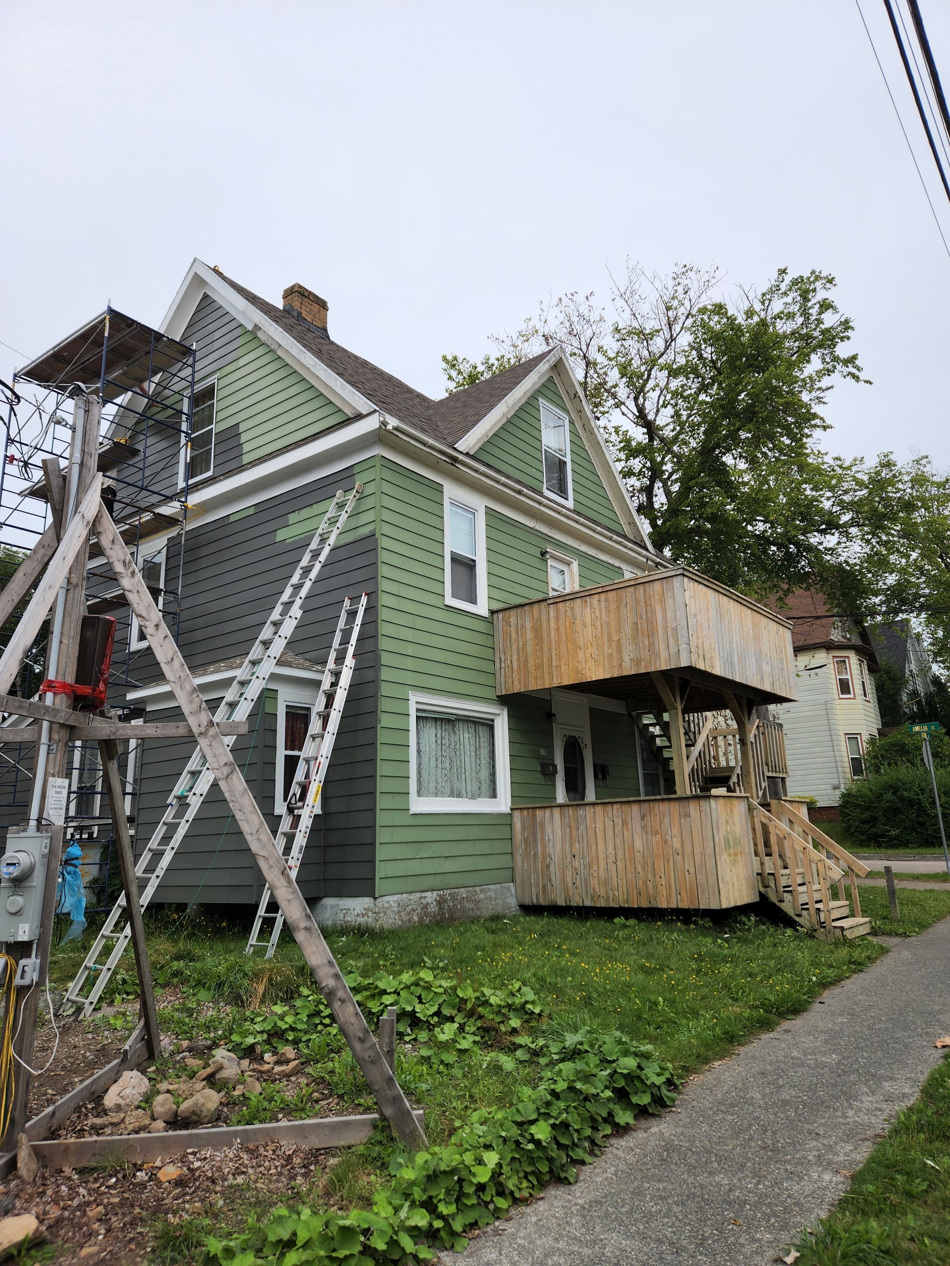 Green house with damaged scaffolding and a wooden porch, beside a sidewalk and tree after storm damage