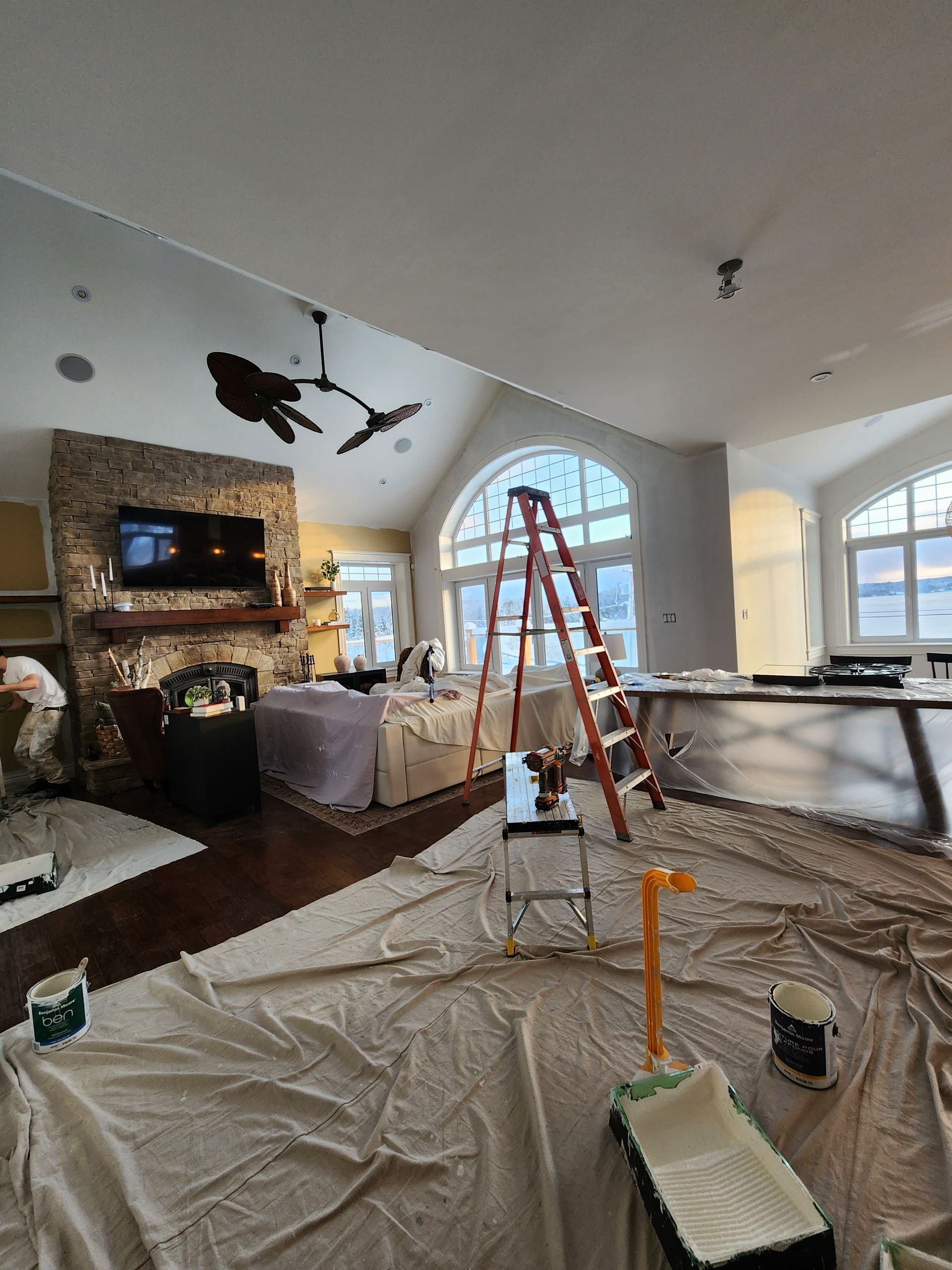 Living room under renovation with tarps, ladders, paint supplies, and exposed ceiling light fixtures