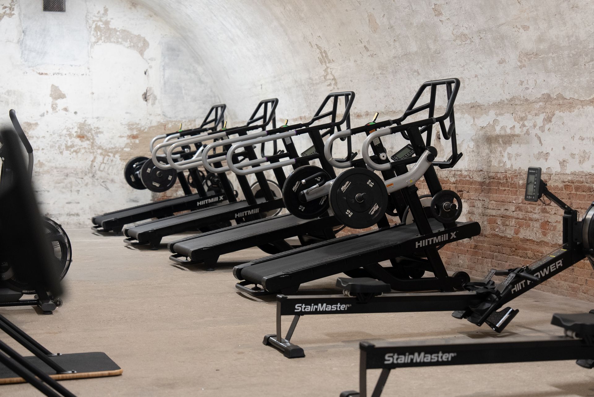 Gym machines and sleds lined up in a worn training room with a concrete wall