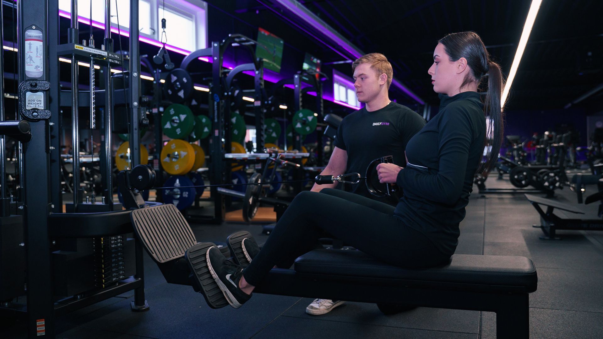 Twee mensen gebruiken roeimachines in een sportschool met neonverlichting, spiegels en andere fitnessapparatuur.