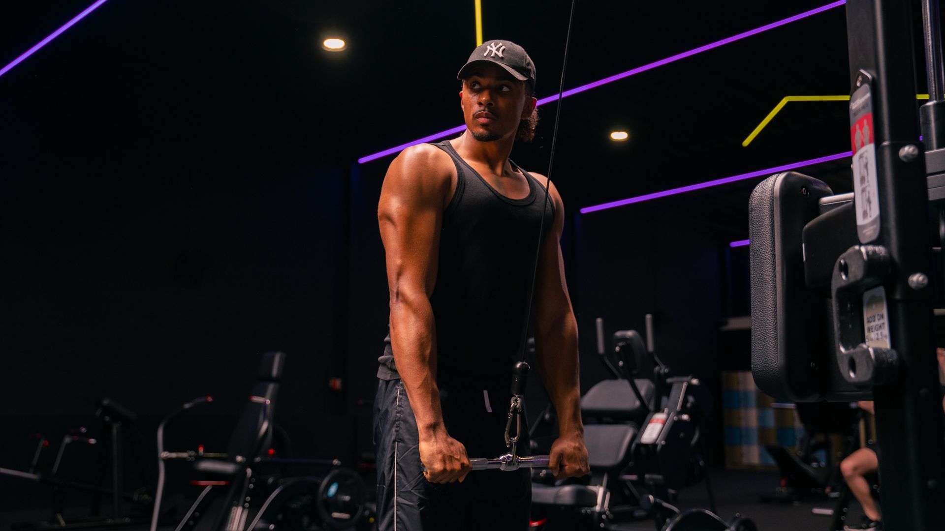 Gym bench press station in a dimly lit fitness room with equipment and sunlight from a window