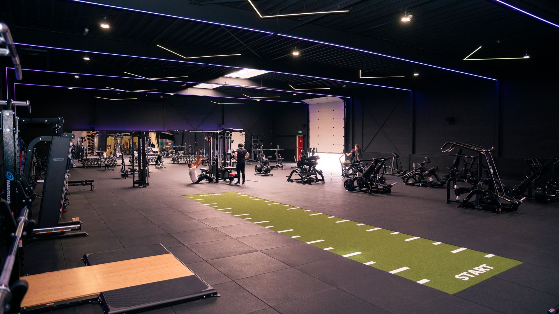 Empty gym with exercise machines, black flooring, and a green turf strip under bright ceiling lights