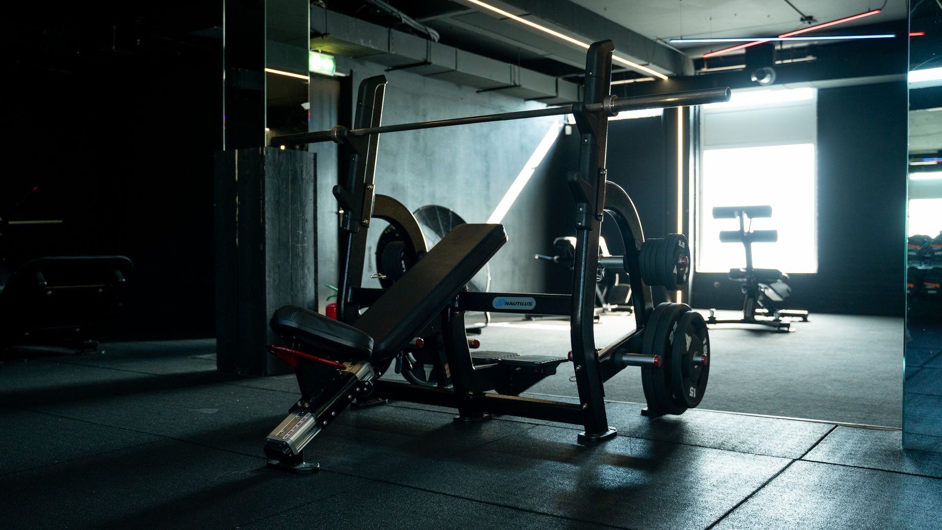A dim gym with a weight bench and barbell rack near a bright window.