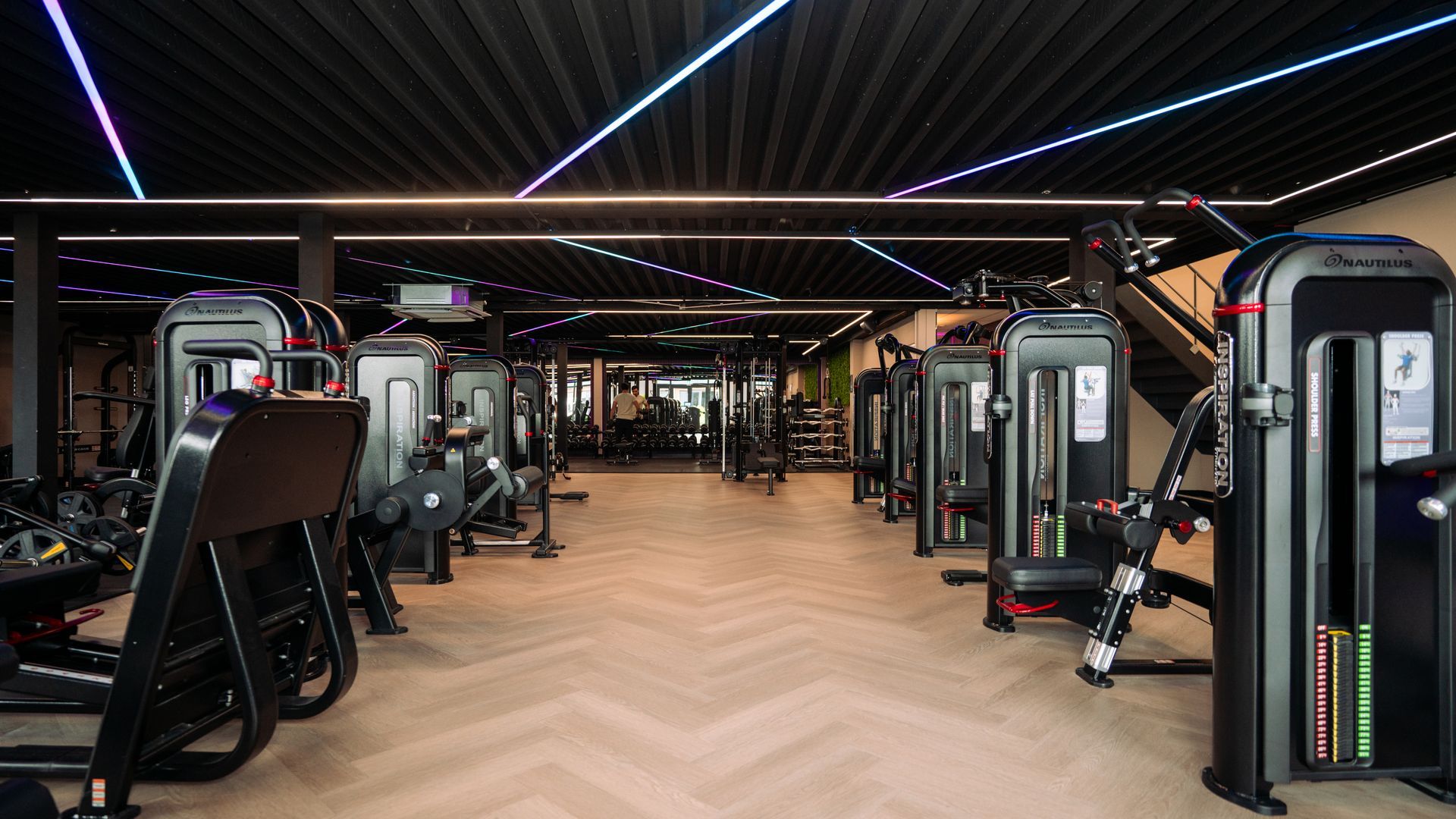 Modern gym interior with rows of exercise machines, wood flooring, and neon ceiling lights