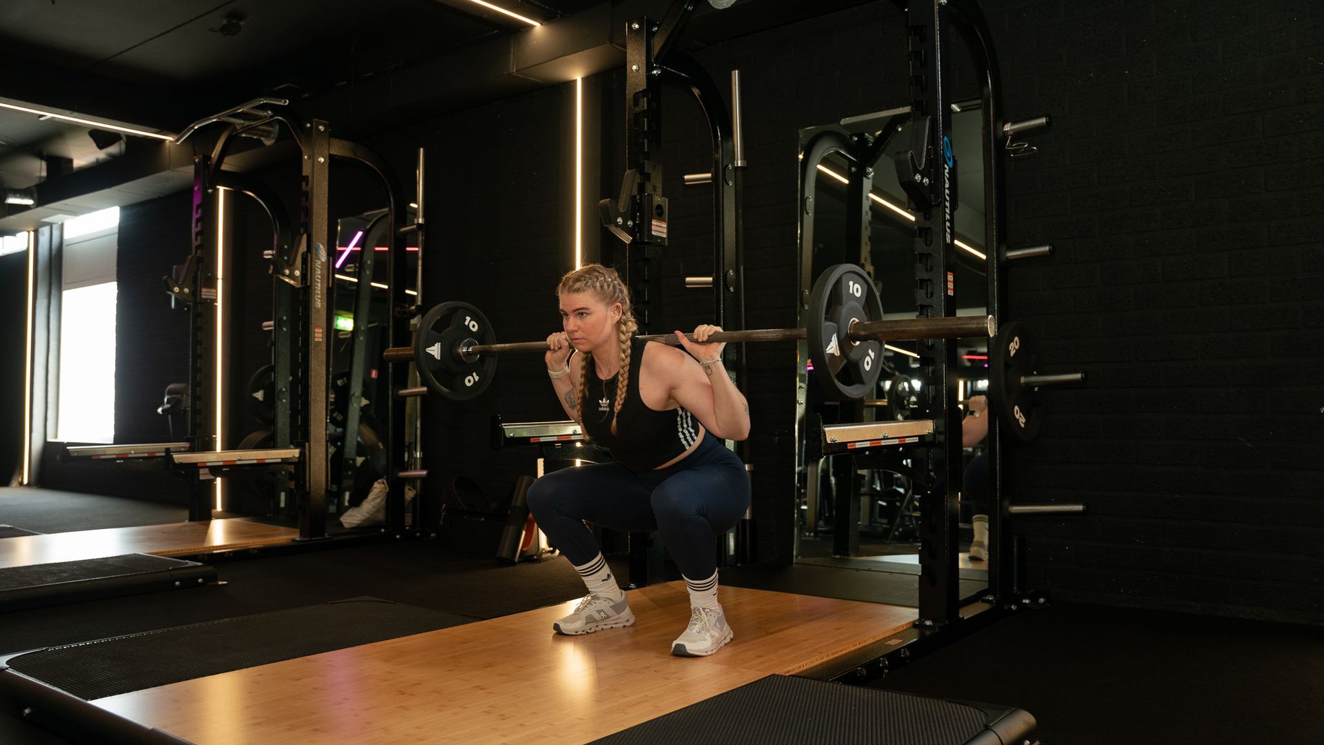 Person squatting with a barbell in a dark gym, wearing black workout clothes and white sneakers.