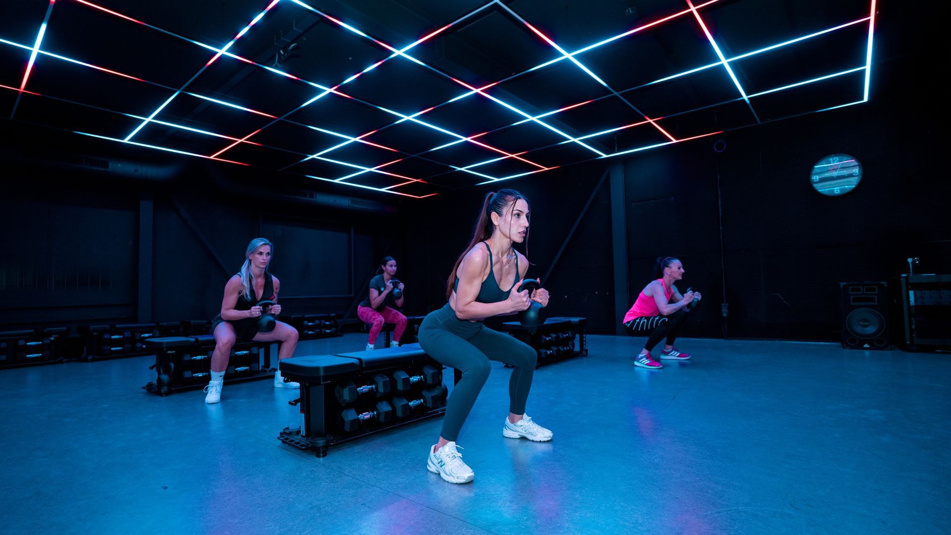 People exercising on benches in a neon-lit fitness studio with blue floor and glowing ceiling lights