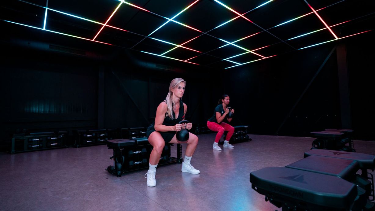 Two people sitting on benches in a neon-lit dance studio with mirrored walls and colorful ceiling lights