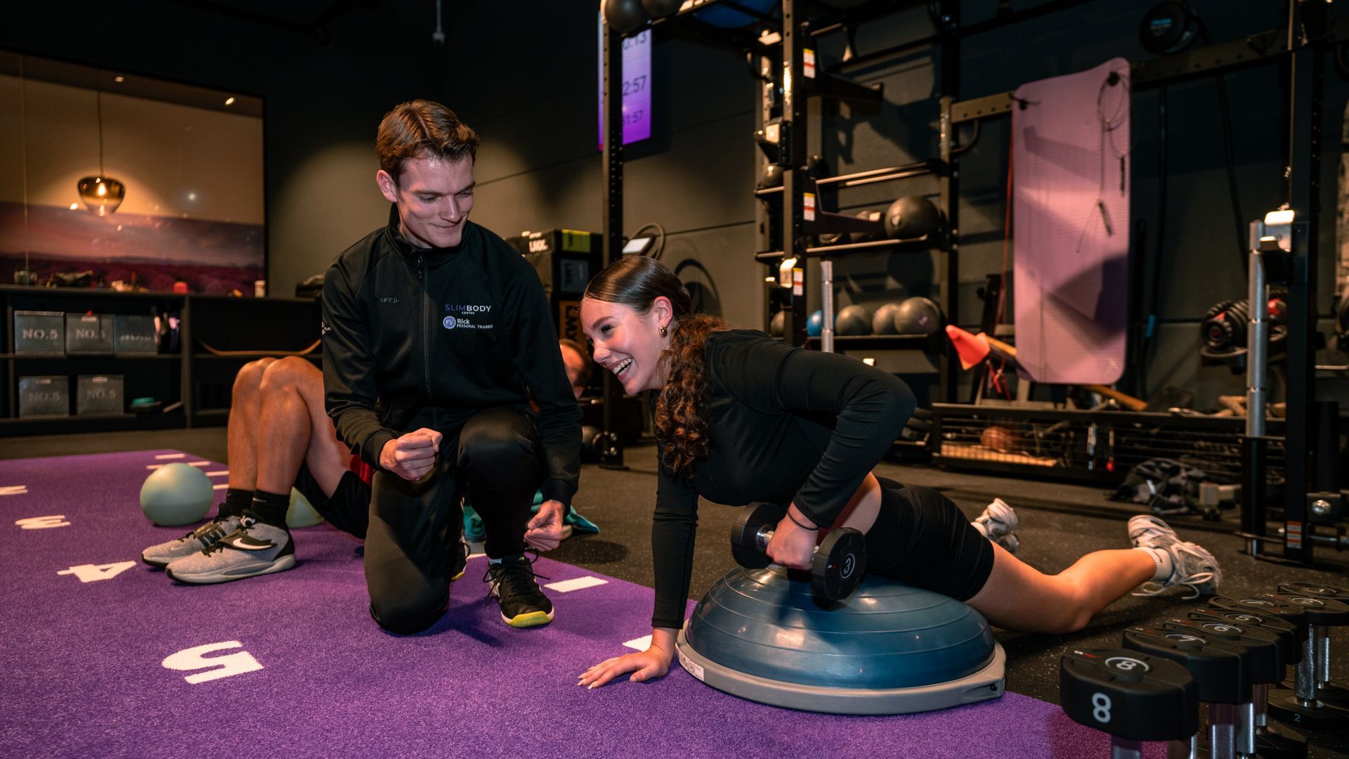 Two people exercising on BOSU balance trainers in a gym studio, one guided by a trainer