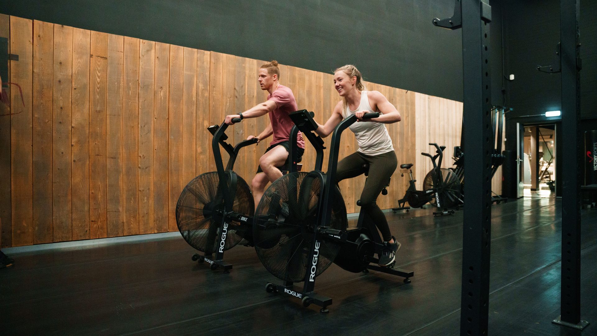 Two people riding stationary exercise bikes in a dim indoor studio with a wooden wall backdrop
