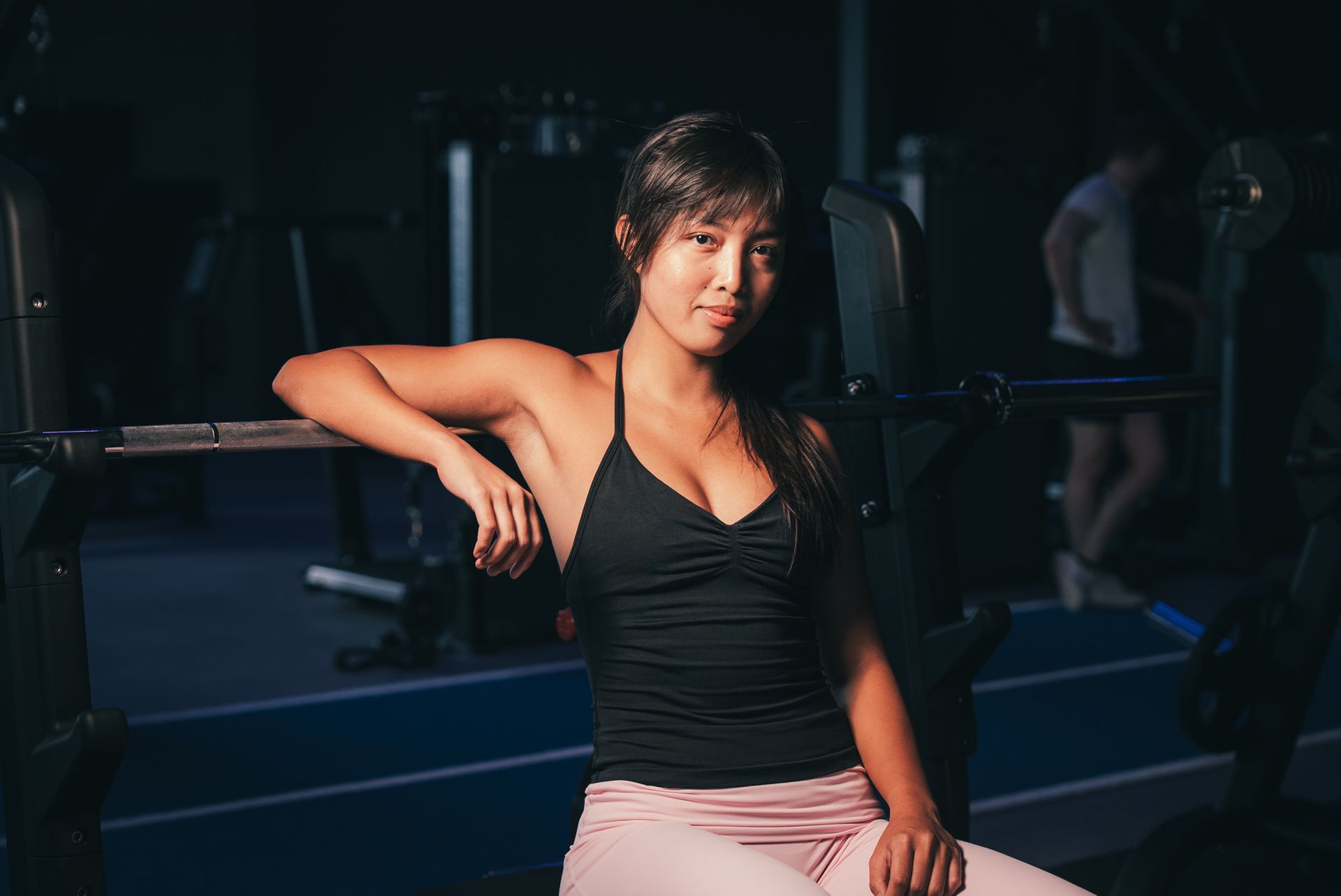 Woman seated in a gym, leaning on a weight bench in a black tank top and pink pants