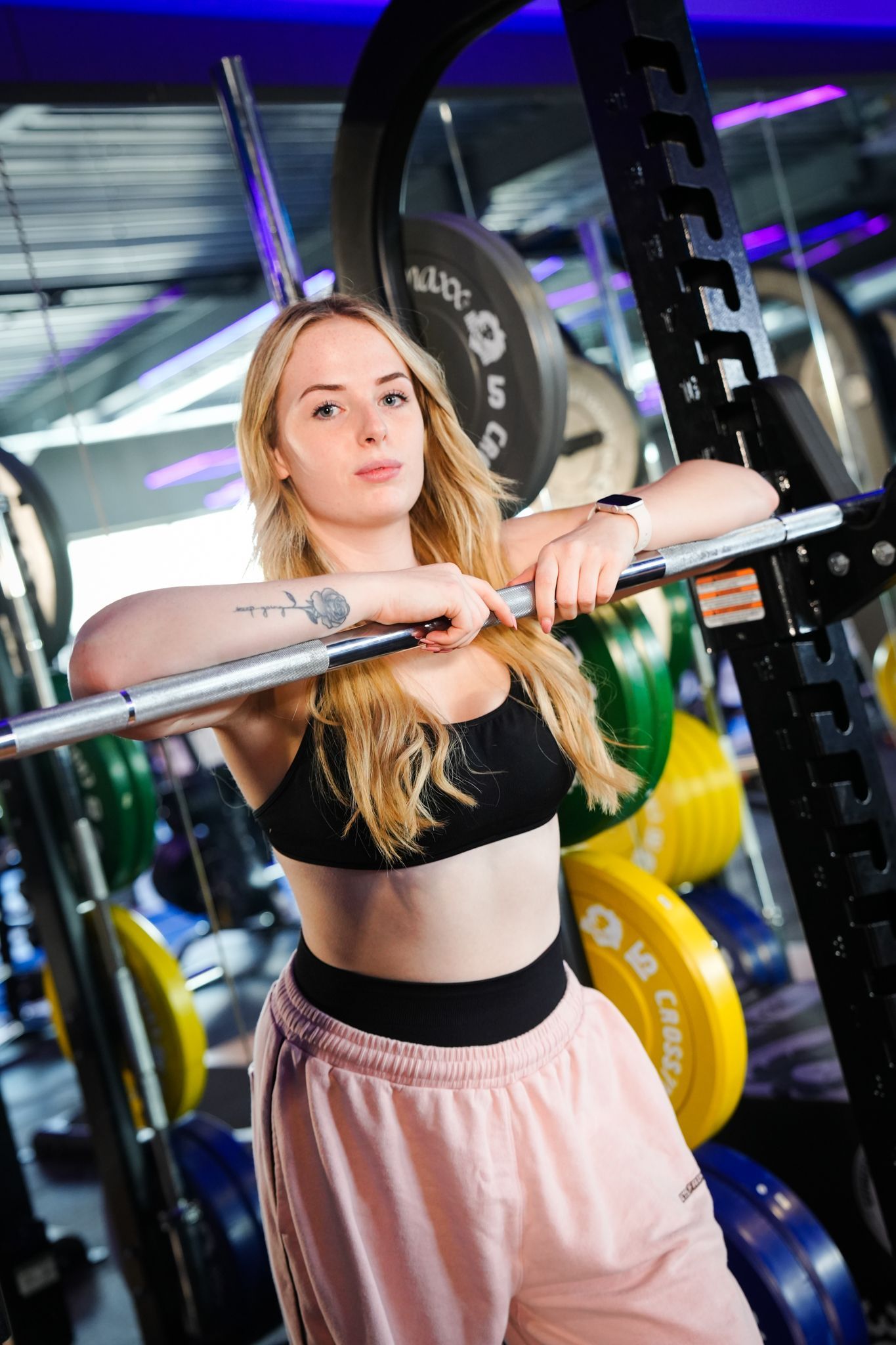 Vrouw in een sportschool, leunend tegen een halterrek, gekleed in een zwarte sportbeha en een roze broek.