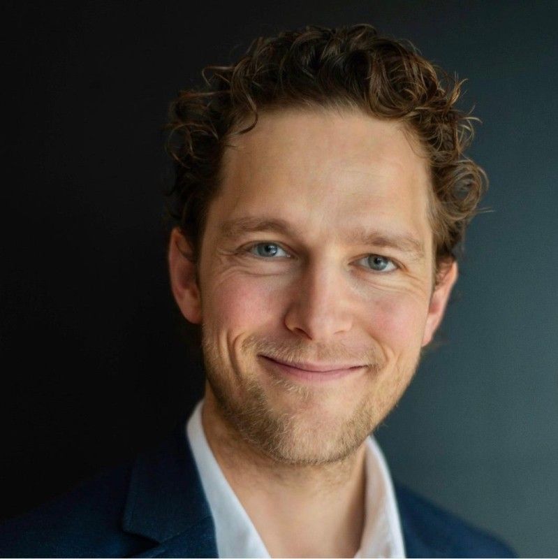 Portret van een lachende man in een donkerblauwe blazer tegen een donkere achtergrond.